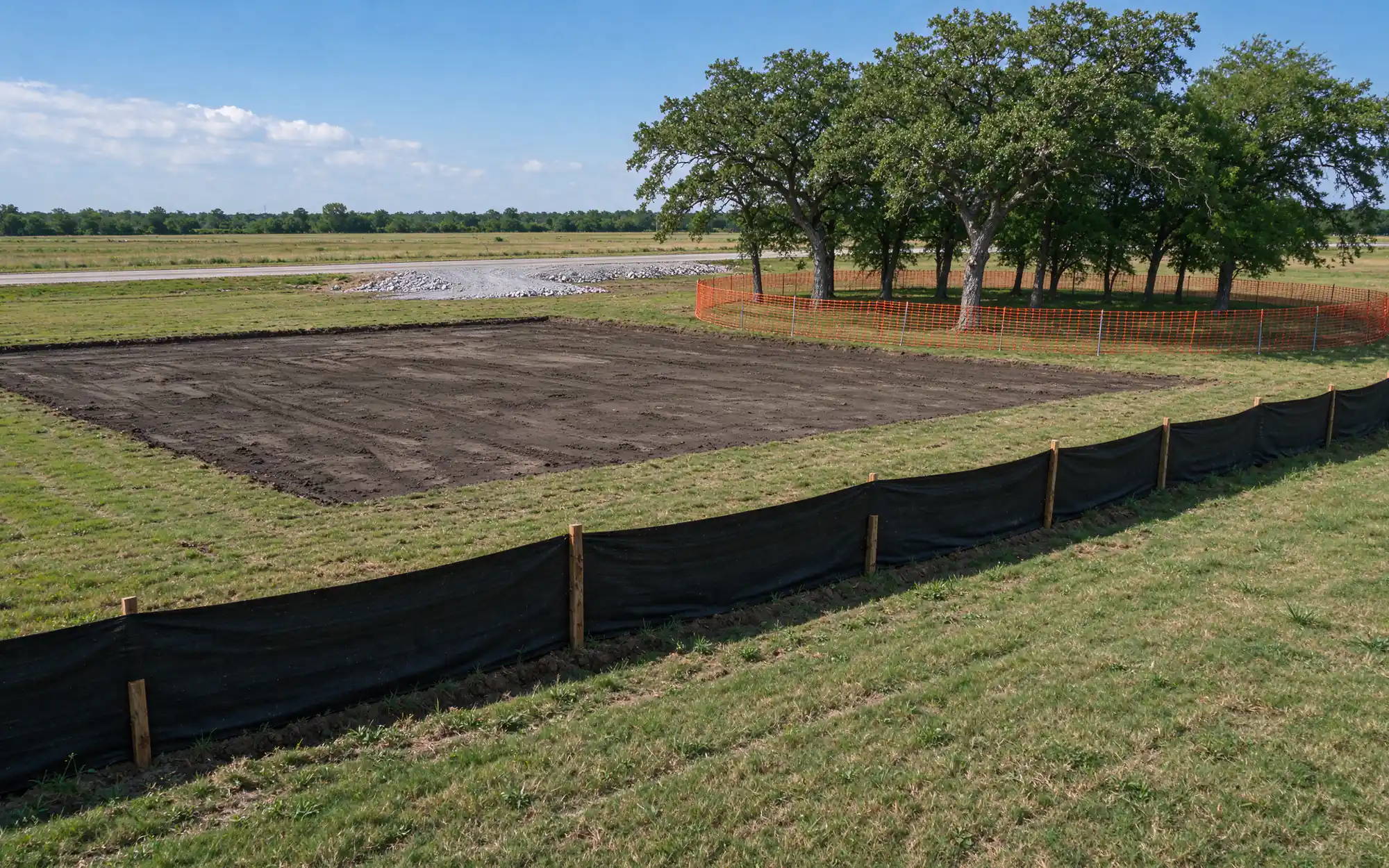 Commercial tract on the North Texas prairie edge with a cleared building footprint of bare dark clay, a line of black silt fence along the low side and a stand of preserved trees behind orange protective fencing